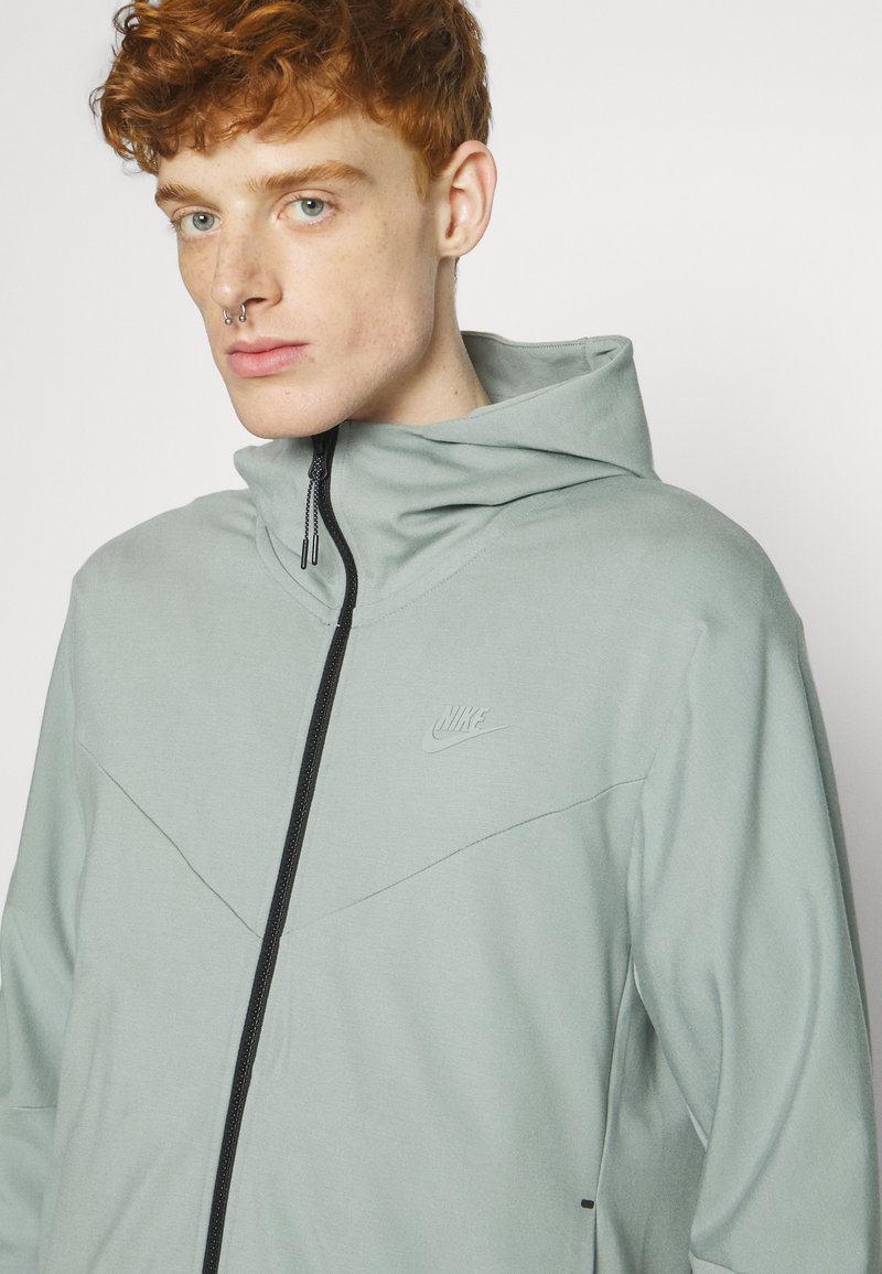 Un jeune avec des cheveux bouclés roux porte un sweatshirt zippé gris clair de Nike avec un col haut, tourné vers l'avant contre un fond uni.