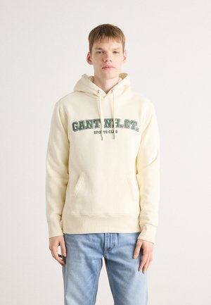 Felpa cream con testo verde "GANT N.H.C.T. SPORTS CLUB", tasca a marsupio frontale, cappuccio con cordoncino, polsini e fondo a costine. Indossata con jeans azzurri chiari.