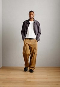 Man walking indoors wearing a gray jacket, white shirt, tan cargo pants, and black shoes on a wooden floor with plain gray walls.