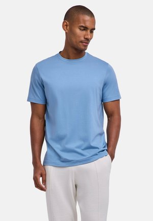 Uomo che indossa una t-shirt a maniche corte azzurro chiaro e pantaloni beige chiaro, in piedi con una mano in tasca, che guarda verso il basso.