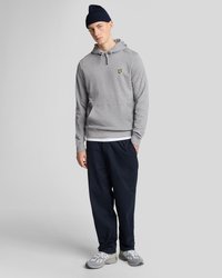 Young man wearing a gray hoodie with a yellow logo, navy pants, gray sneakers, and a navy knit beanie, standing on a plain gray background.