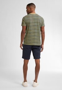 Chemise à manches courtes pour homme à rayures vertes et jaunes avec un col rond, assortie à un short marine foncé et des chaussures de couleur claire.