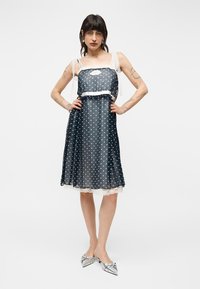 Femme aux cheveux foncés et tatouages portant une robe mi-longue noire transparente à pois avec un bord en dentelle blanche et des chaussures argentées à bouts pointus.