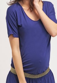 T-paita tummansinisestä kankaasta, jossa on lyhyet hihat, pyöreä kaula-aukko ja istuva vyötärö, jota korostaa kultainen metallinen nauha. Silkkinen, pehmeä kangas.