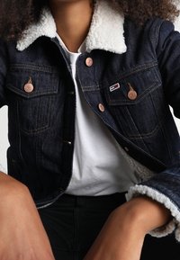 Denim jacket with a sherpa collar, copper buttons, and front pockets; dark blue color with visible stitching and logo detail.