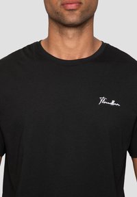 T-shirt en coton noir avec un col rond, des manches courtes et un logo brodé blanc sur la poitrine gauche. Texture lisse.