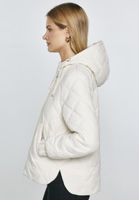 Puchowa kurtka pikowana w odcieniu off-white, z kapturem, kieszeniami bocznymi, zapięciem na sznurki oraz lekko zaokrąglonym dołem. Gładka struktura.