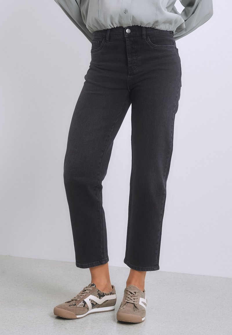 Frau trägt schwarze High-Waist-Jeans und beige Schnürsneaker mit weißem und Schlangenmuster, steht vor einem schlichten hellen Hintergrund.