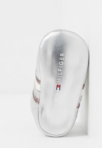 Silberner Sneaker mit glattem Metallic-Finish, ausgestattet mit braunen und weißen Streifen, Logo auf der Sohle und abgerundetem Zehenbereich.
