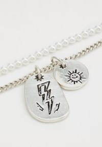 Silverhalsband med två berlocker: en oval med ett blixtdesign och en rund med en sol och ett smiley-ansikte. Pärlad kedjesmyckning.
