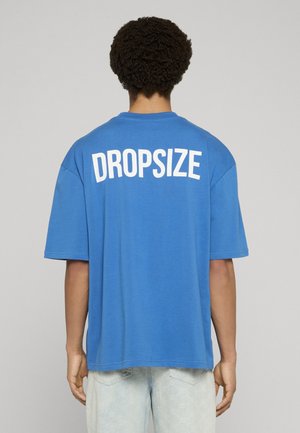 Persona con capelli corti e ricci che indossa una maglietta blu larga con la scritta "DROPSIZE" in bianco sul retro e jeans chiari, in piedi su uno sfondo semplice.