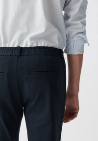 Marineblauwe broek gemaakt van gladde stof, met een tailleband en twee achterzakken, gecombineerd met een lichtblauw gestreept shirt dat bovenuit komt.