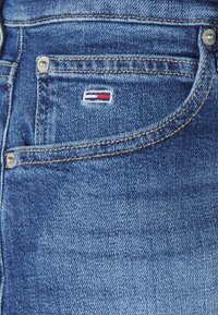 Jeans in denim con un lavaggio blu sbiadito, cuciture visibili e un piccolo logo ricamato con strisce rosse, bianche e blu sulla tasca.