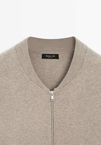 Cardigan lavorato a maglia beige con colletto a costine e chiusura con zip. Presenta una superficie liscia e un'etichetta del marchio all'interno del collo.