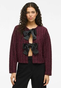 Burgundy texturerad cardigan med tre svarta satinfjädrar vid halsringningen; kort skuren och l̊anga ärmar. Bärs över svarta byxor.