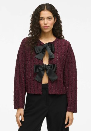 Cardigan texturé bordeaux avec trois nœuds en satin noir au niveau du col ; coupe cropped et manches longues. Porté sur un pantalon noir.