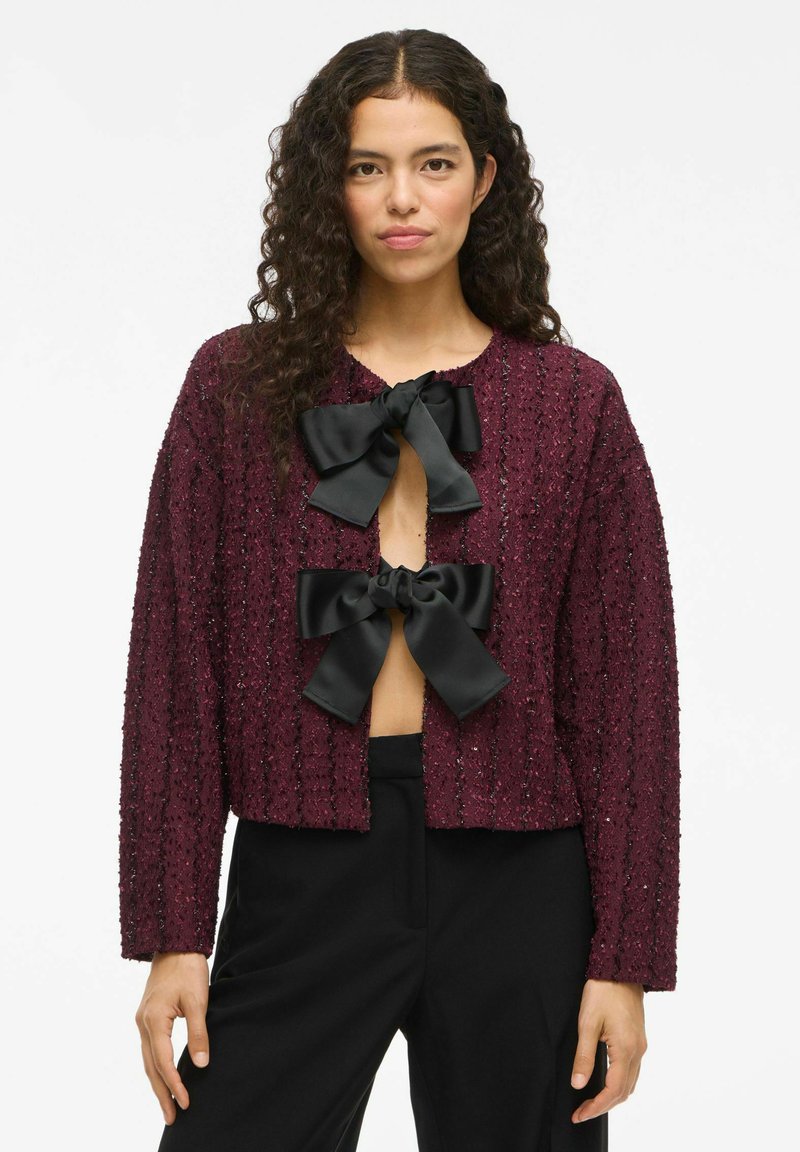 Burgundy texturerad cardigan med tre svarta satinfjädrar vid halsringningen; kort skuren och l̊anga ärmar. Bärs över svarta byxor.