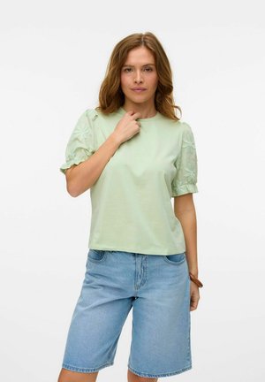 T-shirt verde chiaro a maniche corte con ricami floreali sulle maniche, abbinato a pantaloni corti in denim blu chiaro. Tessuto morbido, vestibilità rilassata.