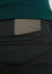 Close-up van een donkere visgraatstof broekzak met een gestikt leren label met de tekst "CHASIN'" onder de zoom van een groene gebreide trui.