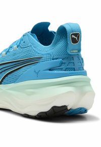 Zapatilla deportiva azul claro con parte superior de punto texturizada, acentos negros, suela verde menta y un distintivo logo en la parte trasera.