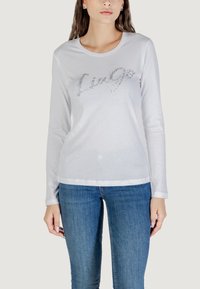 Maglietta bianca a maniche lunghe con scollatura rotonda, caratterizzata da una scritta in glitter argento sulla parte anteriore, abbinata a jeans blu.
