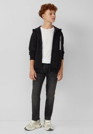 Zwarte zip-up hoodie met een witte grafisch ontwerp aan de linkerkant, gecombineerd met een witte t-shirt, donkere jeans en lichtkleurige sneakers.