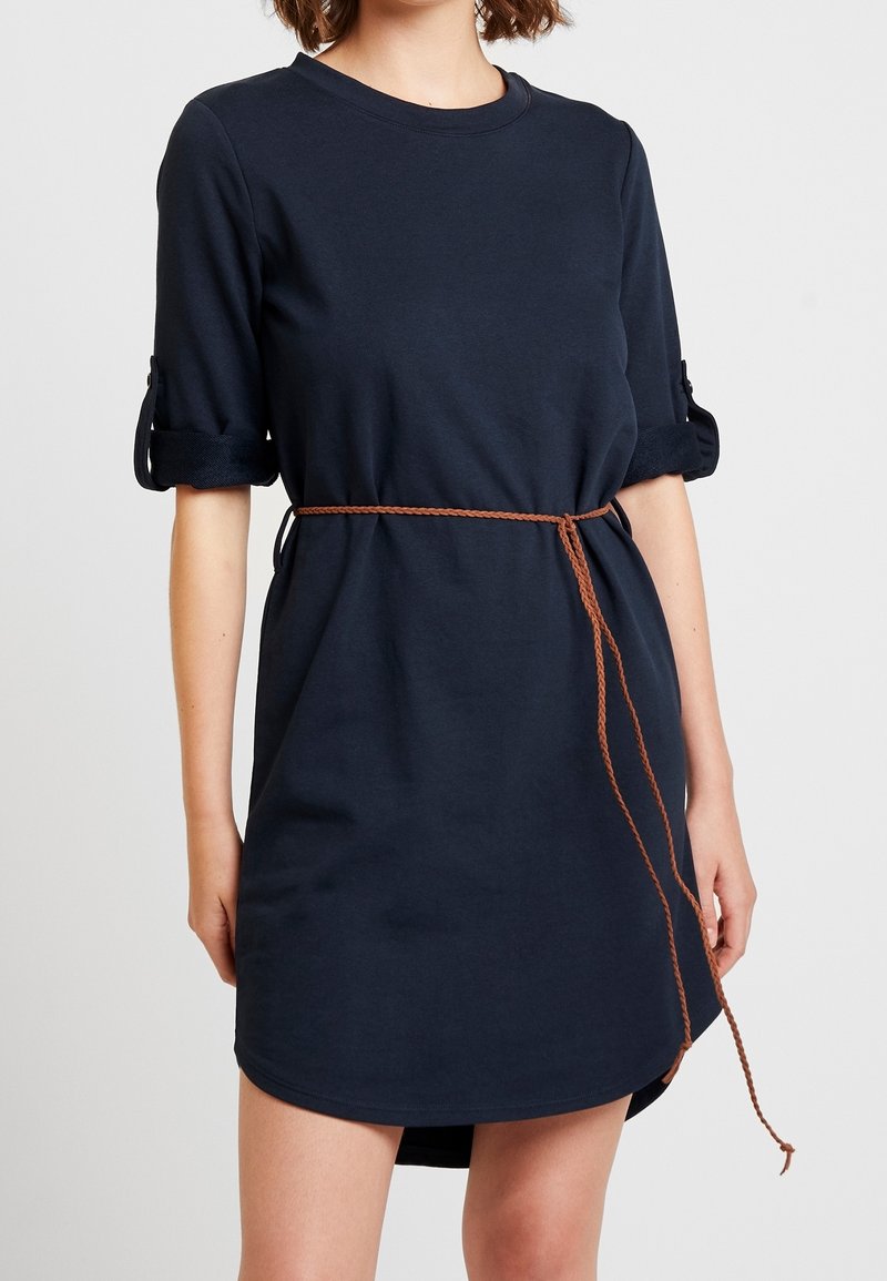 Femme portant une robe bleu marine à manches courtes avec des manches retroussées et une fine ceinture tressée marron nouée à la taille.