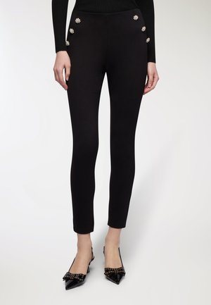 Donna che indossa pantaloni neri slim-fit con sei bottoni decorativi in perla vicino alla vita e scarpe nere con punta e dettagli a fiocco.