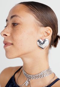 Pendiente plateado en forma de corazón, con superficie lisa y de gran tamaño. Llevado con un gargantilla de plata en capas y una blusa oscura. Piel con pecas visible.