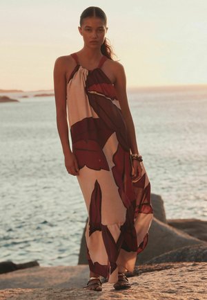 Frau steht am felsigen Ufer bei Sonnenuntergang, trägt ein ärmelloses, luftiges Maxikleid mit Blumenmuster und Sandalen, mit Armband am Handgelenk.