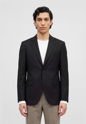 Un homme pose avec une veste noire à deux boutons sur une chemise blanche et un pantalon gris, devant un fond clair uni.