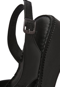 Gros plan sur une sandale compensée en cuir noir avec une bride de cheville réglable et une boucle en métal.