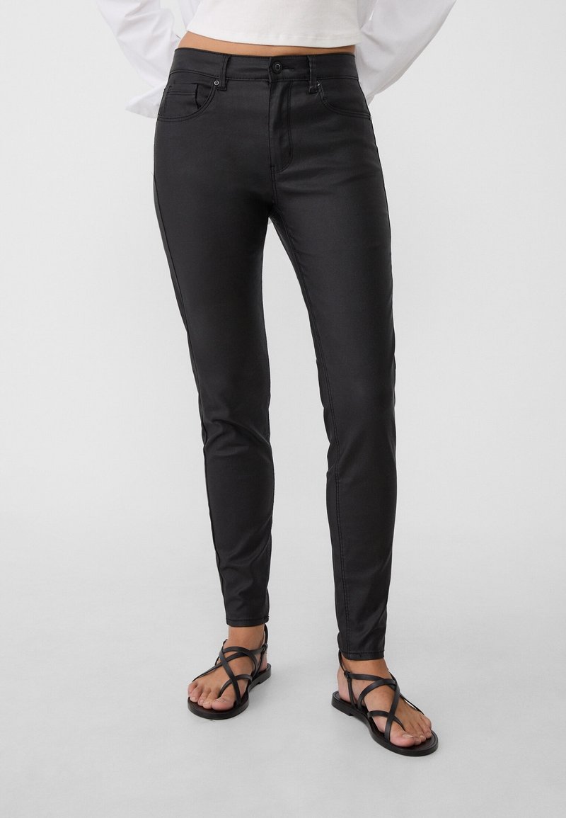 Zwarte slim-fit jeans met een soepele structuur, vijf zakken en riemlussen, gedragen met zwarte sandalen met banden op blote voeten.