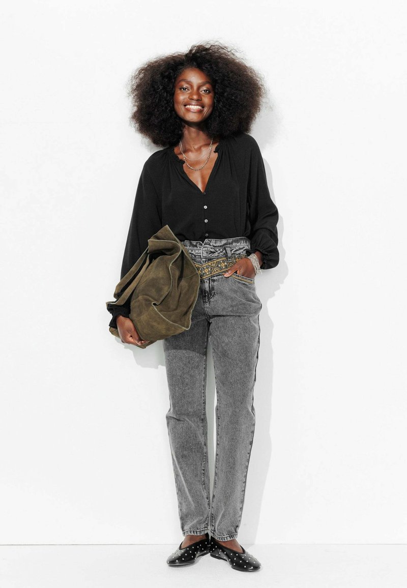 Chemisier noir à boutons, jeans gris taille haute avec une ceinture à motifs, grand sac vert olive, et chaussures noires avec des pois blancs.