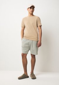 Camiseta beige de manga corta, pantalones cortos de rayas verdes y blancas, y zapatos grises tipo slip-on, acompañados de una gorra de color claro. Conjunto casual.