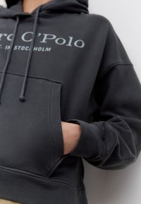 Zwarte hoodie met een voorzak, trekkoord in de capuchon en het logo "Marc O'Polo" in lichtblauw. Zachte textuur met geribbelde boorden.
