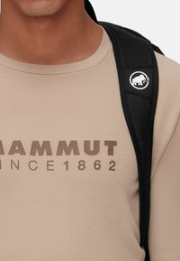 Beige overhemd met lange mouwen met een bruine "Mammut" logo en jaartal, gecombineerd met een zwarte rugzakriem met een wit olifanticoon.