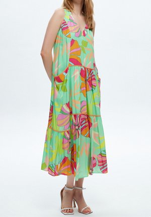 Kobieta w bezrękawnej, luźnej sukience midi z dużym, kolorowym kwiatowym wzorem i białych sandałach na wysokim obcasie z paskiem na kostce.