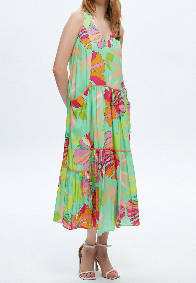 Femme portant une robe midi sans manches, ample, avec un grand imprimé floral coloré et des sandales blanches à talons hauts avec bride à la cheville.