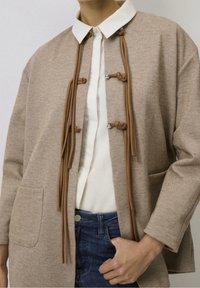 Beżowa kurtka o teksturze i strukturalnym designie, z białym kołnierzykiem, brązowymi akcentami w formie węzła oraz przednimi kieszeniami. Stylizowana na białej bluzce.