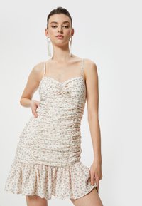Vestido mini blanco con diseño arrugado, tirantes finos, escote corazón y dobladillo con volantes. Acentuado con pequeños patrones de flores rosas.