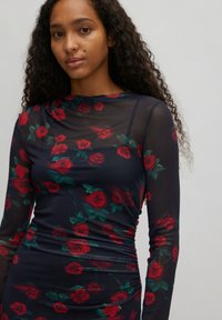 Blumenkleid mit langen Ärmeln aus transparentem schwarzen Stoff, verziert mit roten Rosen und grünen Blättern, mit gerafften Details an der Taille.