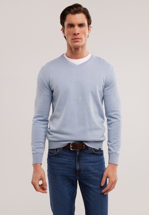 Lichtblauwe v-hals trui van zacht materiaal, gedragen over een wit T-shirt, gecombineerd met donkerblauwe jeans en een bruine riem.