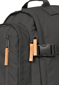 Sac à dos gris foncé en tissu texturé. Équipé de tirettes de fermeture en cuir orange et d'une fermeture à boucle noire robuste.