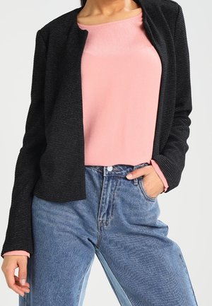 Femme portant un cardigan noir côtelé ouvert sur un haut rose clair et un jean bleu taille haute avec une main dans la poche.
