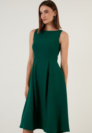 Robe verte sans manches avec un corsage ajusté, une jupe évasée et des détails plissés. Fabriquée en tissu lisse avec un léger éclat.