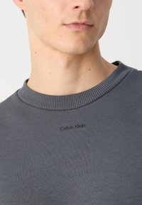 Šedá mikina s okrúhlym výstrihom z jemného materiálu, s rebrovaným golierom a decentným embosovaným logom Calvin Klein na hrudi.
