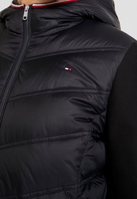 Zwarte pufferjack met capuchon, met een gewatteerde textuur, ritssluiting en een klein kleurrijk logo op de borst.