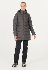 Grå isoleret jakke med lodret quiltning, lynlås foran, justerbar hætte og sidelommer, båret over sorte bukser og vandrestøvler.