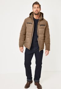Brown puffer jacket with a hood, zippered chest pockets, quilted texture, worn over a navy sweater and dark jeans, paired with brown boots.